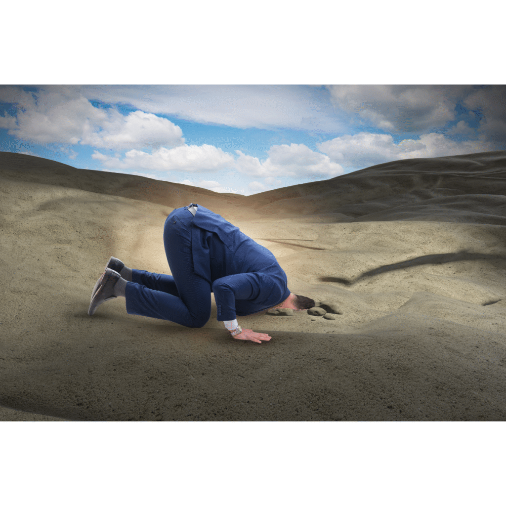 Businessman in a suit kneeling on the ground with his head buried in the sand, symbolizing avoidance or denial of important issues.