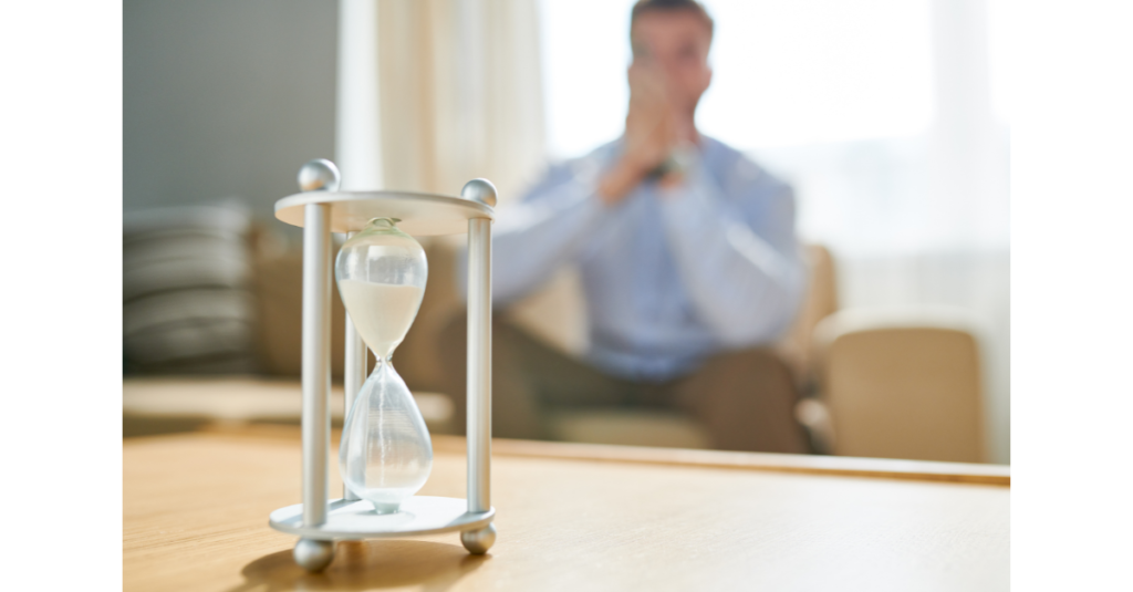Hourglass on a table in the foreground while a man sits in the background with hands clasped, appearing thoughtful as time passes